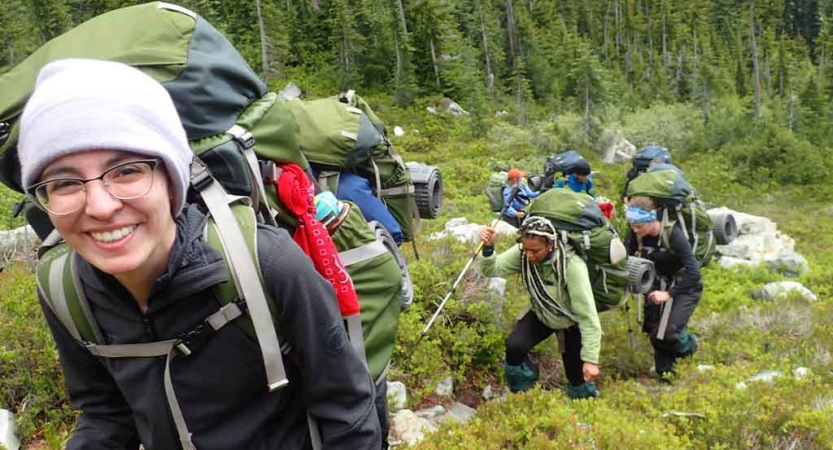a person wearing a backpack smiles at the camera, while others follow behind them on a trail